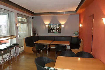 Innenraum einer Bar mit Holztischen, schwarzen Stühlen, drei Barhockern und einer Eckbank. Fenster mit Jalousien und Wandbild mit Schriftzug „Home Sweet Home“ auf einer grauen Wand. Rechts eine orangefarbene Wand mit angebrachten Leuchten.