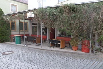 Außenbereich einer Kneipe mit roten Sitzbänken, Holztischen und schwarzen Stühlen. Vor dem Gebäude ein grünes Fass als Tisch auf einem gepflastertem Platz aufgestellt. Am Dach entlang verläuft ein grünes Gewächs, das auf einer Seite fast bis zu Boden runterhängt. Die Überdachung ist mit rot-gelben LED-Streifen beleuchtet.