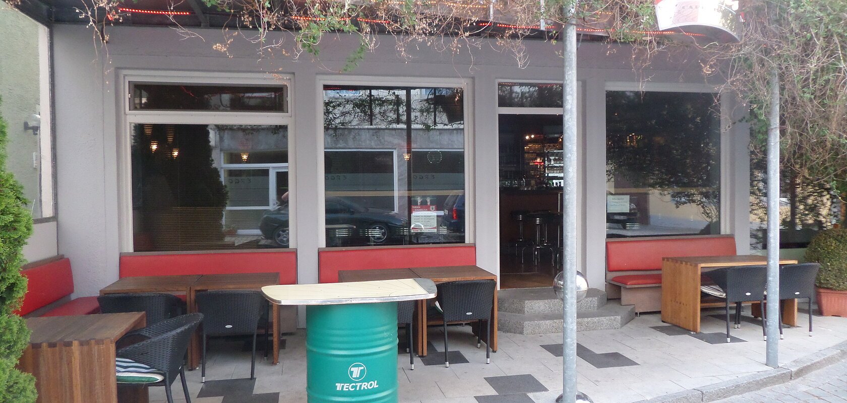 Außenbereich einer Kneipe mit roten Sitzbänken, Holztischen und schwarzen Stühlen. Vor dem Gebäude ein grünes Fass als Tisch auf einem gepflastertem Platz aufgestellt. Am Dach entlang verläuft ein grünes Gewächs. Die Überdachung ist mit rot-gelben LED-Streifen beleuchtet.