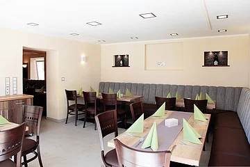 Blick in einen hellen Gastraum einer Wirtschaft mit Holztischen, Holzstühlen,  grau-braunen Polstersitzbänken an den Wänden, grünen Servietten und Besteck. In den beige gestrichenen Wänden sind drei dekorative beleuchtete Ausfräsungen.