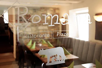 Fensterglas einer Pizzeria mit weißem Schriftzug „Romana Ristorante Pizzeria“ und Logo, dahinter unscharf zu sehen gedeckte Tische mit grünen Servietten und einer Steinwand.