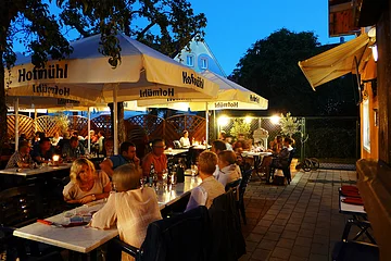 Abendliche Außengastronomie mit Gästen an Tischen unter großen Sonnenschirmen und Beleuchtung auf gepflastertem Boden.