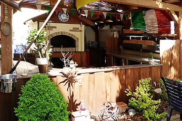 Außenküche mit Holztheke, Pizzaofen, italienischer Flagge und Pflanzen im Gartenbereich. Recht im Bild ein grauer Stuhl zu sehen.
