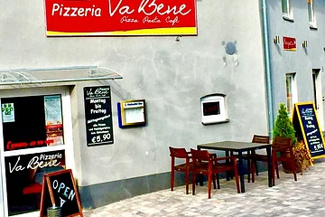Außenansicht einer grau gestrichenen Pizzeria mit Tischen, Stühlen und mehreren Schildern mit Öffnungszeiten und Angeboten vor dem Gebäude. Zwei bepflanzte Töpfe stehen auf dem gepflastertem Platz.
