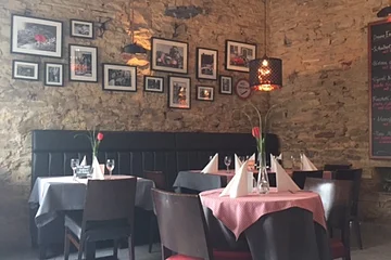 Restaurant mit beleuchteter Steinwand, mehreren Tischen mit weißen und roten Tischdecken, Stühlen und Wandbildern. Rechts im Bild an der Wand eine Tafel mit den aktuellen Gerichten angebracht.