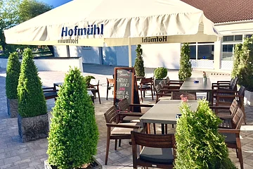 Außenbereich einer Pizzeria mit Tischen, Stühlen und einem Sonnenschirm umgeben von bepflanzten Kübeln auf gepflastertem Boden. Im Hintergrund ein Haus, Bäume und blauer Himmel zu sehen.