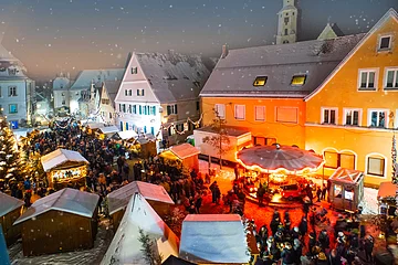 Der Monheimer Weihnachtsmarkt mit zahlreichen Besuchern, festlich beleuchteten Ständen und Kinderkarussell bei Schnee in historischen Altstadt von Monheim.