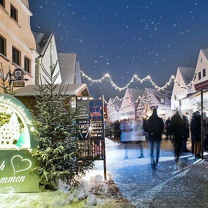 Monheimer Weihnachtsmarkt mit Verkaufsständen, Lichtern und Menschen. Ein beleuchtetes Schild links im Bild mit dem Text: "Monheimer Weihnachtsmarkt - Herzlich Willkommen" umgeben von mehreren Tannenbäumen. Schnee liegt auf dem gepflastertem Platz.