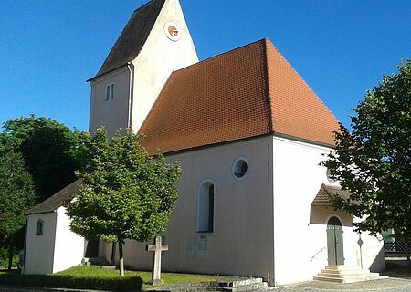 Kirche mit rotem Satteldach und einer Turmuhr umgeben von Bäumen bei klarem Himmel, an einem gepflastertem Weg.