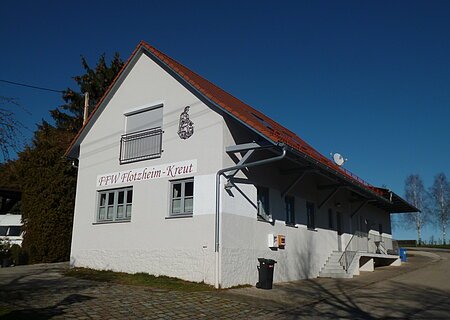 Feuerwehrhaus Flotzheim Feuerwehrhaus mit Aufschrift „FFW Flotzheim-Kreut“ und rotem Ziegeldach bei klarem Himmel. Umgeben von Bäumen, einem gepflastertem Platz und blauem Himmel.