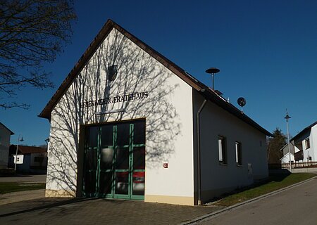 Feuerwehrhaus mit einem großen grünen Tor, Fenstern und braunem Dach an einer Straße. Auf dem weißen Gebäude die Aufschrift "Feuerwehrgerätehaus" und Schatten eines Baumes auf der Fassade, bei klarem Himmel. Links im Eck des Bildes ein Baum zu sehen.