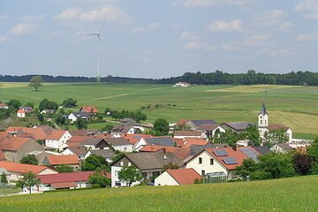 Dorf mit roten Dächern, einer Kirche mit Turm, grünen Wiesen und einem Windrad im Hintergrund, unter blauem Himmel.