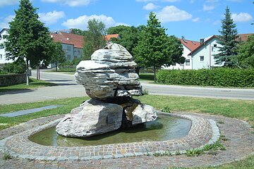 Brunnen mit großen weißen Steinen und Wasser, umgeben von kreisförmig gepflastertem Weg. Im Hintergrund sind Bäume vor Wohnhäuser bei blauem Himmel.