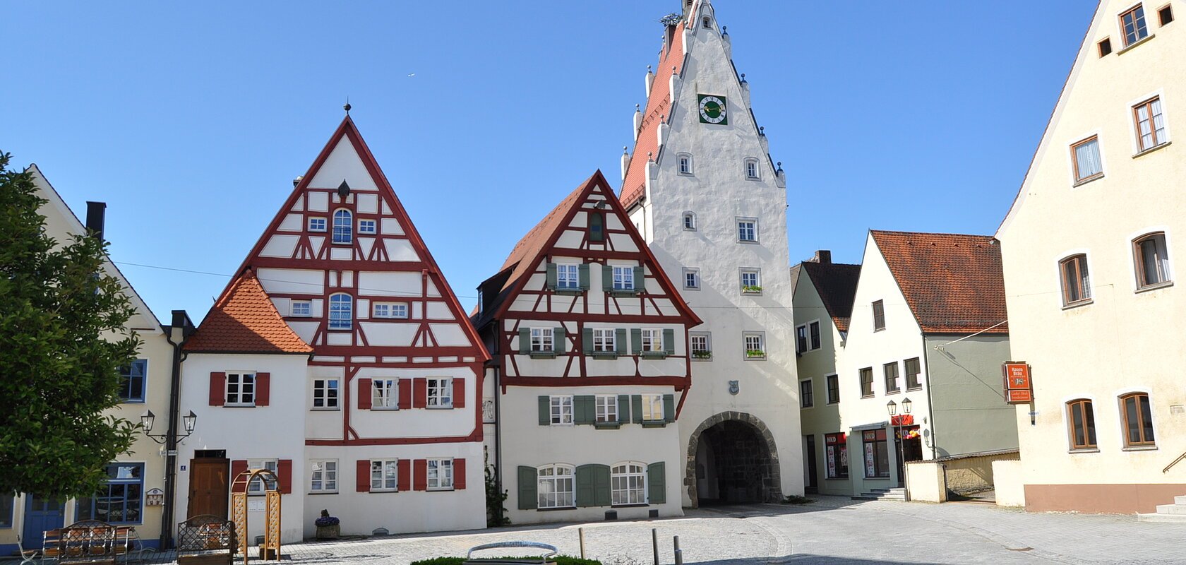 Historische Fachwerkhäuser in einer Innenstadt mit einem weißen Uhrenturm an einem gepflasterten Platz bei klarem Himmel. Im Vordergrund ist ein Brunnen zu sehen, umgeben von drei Sitzbänken.