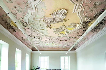 Decke mit barocker Stuck- und Freskoverzierung in einem hellen Raum mit Fenstern und moderner Beleuchtung.