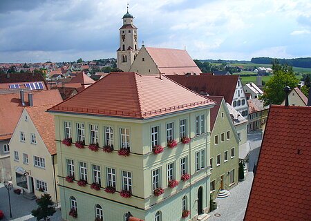 Blick von oben auf eine Innenstadt mit Hauptaugenmerk auf ein historisches grünes Gebäude im Vordergrund, mit roten Dachziegeln und Blumen an den Fenstern.  Im Hintergrund weitere Häuser der Kleinstadt und blauer Himmel. Rechts unten im Eck das rote Dach eines anderen Gebäudes im Vordergrund zu sehen.