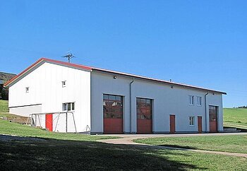 Feuerwehrhaus mit weißen Wänden, roten Türen und großen Roten Toren, vor blauem Himmel und grüner Wiese. Neben dem Gebäude ein Fußballtor.