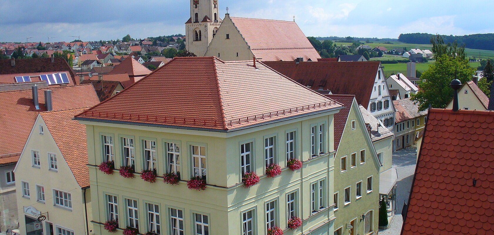 Blick von oben auf eine Innenstadt mit Hauptaugenmerk auf ein historisches grünes Gebäude im Vordergrund, mit roten Dachziegeln und Blumen an den Fenstern.  Im Hintergrund weitere Häuser der Kleinstadt und blauer Himmel. Rechts unten im Eck das rote Dach eines anderen Gebäudes im Vordergrund zu sehen.