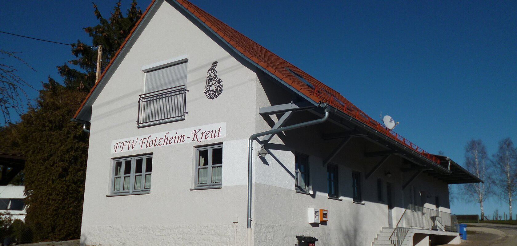 Feuerwehrhaus mit Aufschrift „FFW Flotzheim-Kreut“ und rotem Ziegeldach bei klarem Himmel. Umgeben von Bäumen, einem gepflastertem Platz und blauem Himmel.