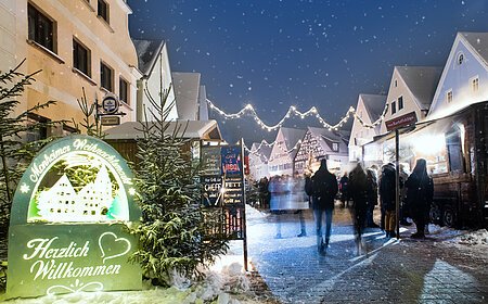 Monheimer Weihnachtsmarkt mit Verkaufsständen, Lichtern und Menschen. Ein beleuchtetes Schild links im Bild mit dem Text: "Monheimer Weihnachtsmarkt - Herzlich Willkommen" umgeben von mehreren Tannenbäumen. Schnee liegt auf dem gepflastertem Platz.