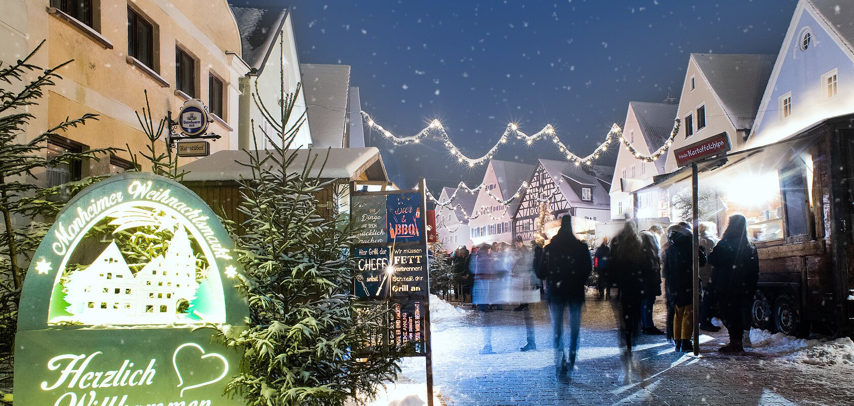 Monheimer Weihnachtsmarkt mit Verkaufsständen, Lichtern und Menschen. Ein beleuchtetes Schild links im Bild mit dem Text: "Monheimer Weihnachtsmarkt - Herzlich Willkommen" umgeben von mehreren Tannenbäumen. Schnee liegt auf dem gepflastertem Platz.