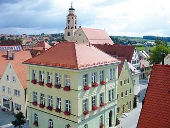 Blick von oben auf eine Innenstadt mit Hauptaugenmerk auf ein historisches grünes Gebäude im Vordergrund, mit roten Dachziegeln und Blumen an den Fenstern.  Im Hintergrund weitere Häuser der Kleinstadt und blauer Himmel. Rechts unten im Eck das rote Dach eines anderen Gebäudes im Vordergrund zu sehen.