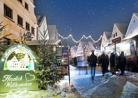 Monheimer Weihnachtsmarkt mit Verkaufsständen, Lichtern und Menschen. Ein beleuchtetes Schild links im Bild mit dem Text: "Monheimer Weihnachtsmarkt - Herzlich Willkommen" umgeben von mehreren Tannenbäumen. Schnee liegt auf dem gepflastertem Platz.