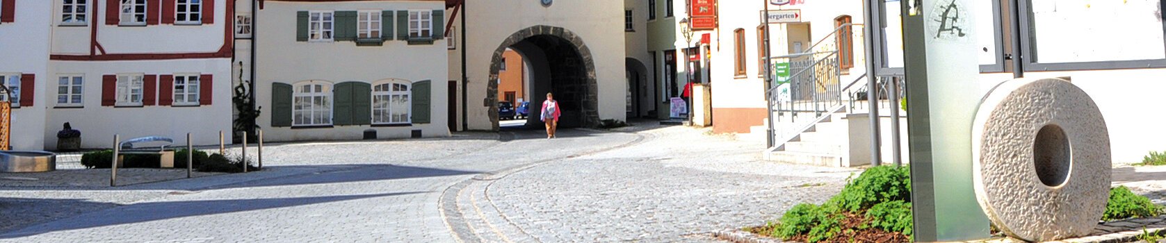 Pflasterstraße mit historischem Torbogen, Fachwerkhäusern und einer Person in der Bildmitte. Rechts im Bild eine Steinskulptur des Buchstabens O mit einer Infotafel dahinter.