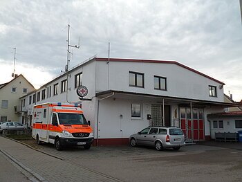 An der Straße gelegenes weißes Gebäude mit Deutschem Roten Kreuz-Schild seitlich am Gebäude, roten Toren und einem Rettungswagen links am Gebäude. Auf dem gepflastertem Platz steht ein grauer Kombi vor dem Gebäude, bei bewölktem Himmel.