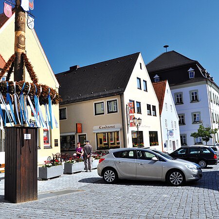 Marktplatz mit einem geschmückten Maibaum im Vordergrund, geparkten Autos an der Straße entlang und historischen Gebäuden bei klarem Himmel in einer Kleinstadt.