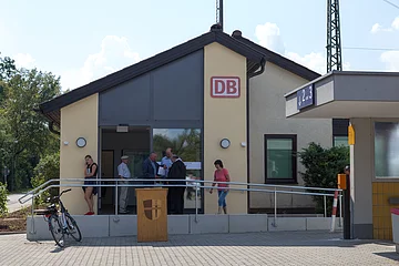 Bahnhofsgebäude in beige-grau von außen mit eingerahmten DB-Logo in rot, sechs Personen vor dem Eingang und ein Fahrrad am Geländer des Aufstiegt. Rechts der unterirdische Gang zu den Gleisen mit eine blauen Schild zu Gleisen 2 und 3.