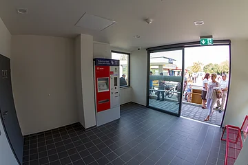Wartehalle am Bahnhof Otting-Weilheim mit einem rot-grauem Fahrkartenautomat an der Wand, einer Glaseingangstür zum Außenbereich, einer roter Sitzbank rechts im Bild und gefliesten Boden. Vor der Tür im Außenbereich mehrere Personen bei Sonnenschein.