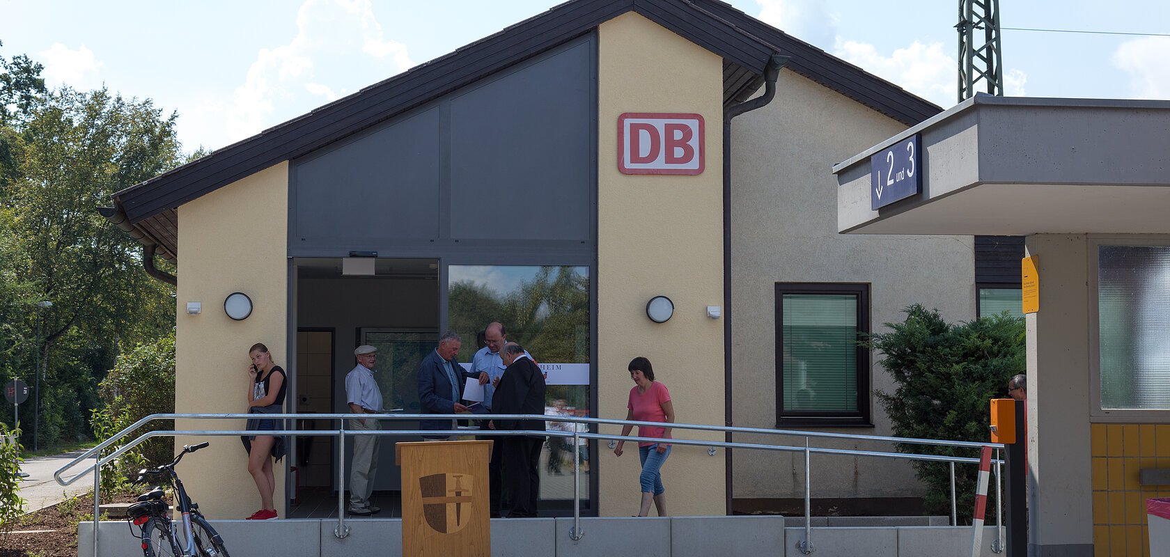 Bahnhofsgebäude in beige-grau von außen mit eingerahmten DB-Logo in rot, sechs Personen vor dem Eingang und ein Fahrrad am Geländer des Aufstiegt. Rechts der unterirdische Gang zu den Gleisen mit eine blauen Schild zu Gleisen 2 und 3.