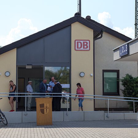 Bahnhofsgebäude in beige-grau von außen mit eingerahmten DB-Logo in rot, sechs Personen vor dem Eingang und ein Fahrrad am Geländer des Aufstiegt. Rechts der unterirdische Gang zu den Gleisen mit eine blauen Schild zu Gleisen 2 und 3.