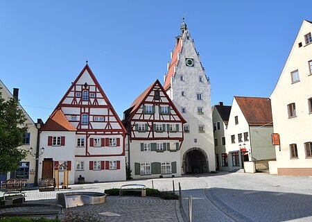 Historische Fachwerkhäuser in einer Innenstadt mit einem weißen Uhrenturm an einem gepflasterten Platz bei klarem Himmel. Im Vordergrund ist ein Brunnen zu sehen, umgeben von drei Sitzbänken.