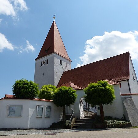 Kirche mit rotem Dach, Turmuhr und umgebender weißer Mauer mit Eingangstor bei blauem Himmel und Bäumen davor.
