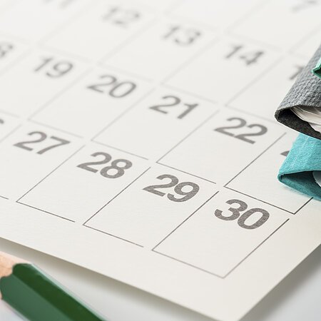Nahaufnahme von einem weißen Kalenderblatt mit schwarzen Datums-Zahlen drauf. Daneben rechts im Bild liegen drei farbige Aktenordner auf dem Kalender und ein grüner Bleistift im Vordergrund.