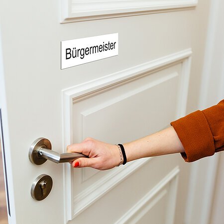 Eine Frauenhand mit roten Nägeln, einem Armband und braunem Oberteil greift an eine Türklinke an weißer Tür, mit Schild „Bürgermeister“. Die Tür zu einem Büro ist halb offen.