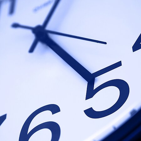 Nahaufnahme einer analogen Uhr mit weißem Hintergrund, schwarzen Zeigern und großen Ziffern auf weißem Zifferblatt.