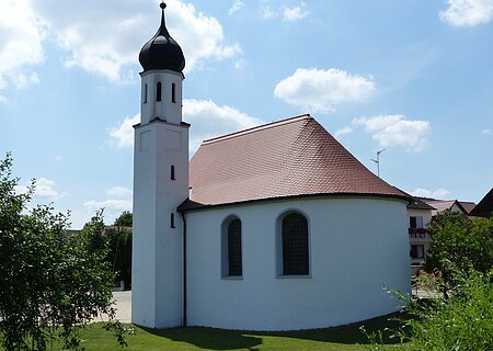 Weißes Kirchgebäude mit abgerundetem Grundriss und kleinem Turm mit schwarzer Haube bei blauem Himmel, umgeben von grünen Bäumen und wiese. Im Hintergrund weitere Häuser.