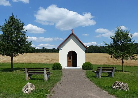 Kleine weiße Kapelle mit rotem Dach an einem gepflastertem Weg mit jeweils einer Bank, einem Baum und einem großen Stein links und rechts am Weg entlang, auf einer Wiese vor Feld und blauem Himmel.
