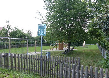Spielplatz mit einem Holzzaun, Schaukeln, Klettergerüst, Rutsche, Bänken und Bäumen auf Grasfläche und teilweise mit Kieselsteinen bei Tageslicht. Außerdem ein am Zaun angebrachtes grünes Schild mit Regeln zur Spielplatzbenutzung und eine Mülltonne aus Metall zu sehen.