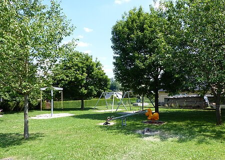 Spielplatz mit Wippen, Schaukeln und einem Sandkasten umgeben von grünen Bäumen auf einer Wiese bei Sonnenschein.