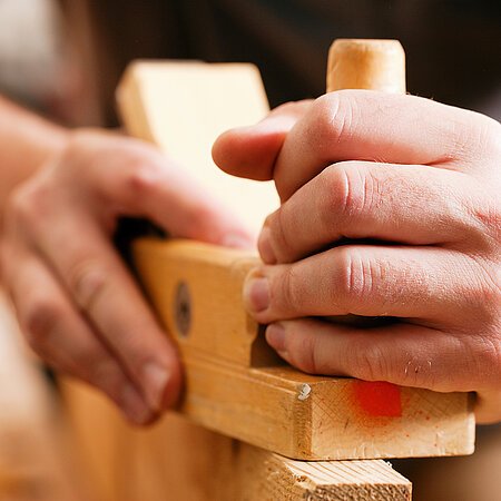 Hände halten eine Hobelmaschine und bearbeiten ein Stück Holz in Nahaufnahme.