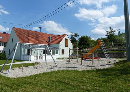 Spielplatz mit einer Schaukel, Klettergerüst, einem Sandkasten mit roter Umrandung und oranger Rutsche, vor einem türkisfarbenen Haus mit rotem Dach, bei blauem Himmel. Umgeben von einer grünen Wiese und Bäumen mit Kieselsteine an den Spielgeräten.
