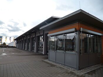 Außenansicht eines modernen grauen Feuerwehrhauses mit Glasfront, flachem Dach, vielen großen Toren und gepflastertem Vorplatz bei bewölktem Himmel.