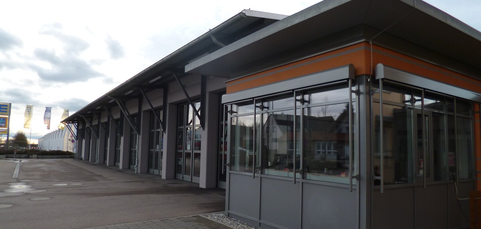 Außenansicht eines modernen grauen Feuerwehrhauses mit Glasfront, flachem Dach, vielen großen Toren und gepflastertem Vorplatz bei bewölktem Himmel.
