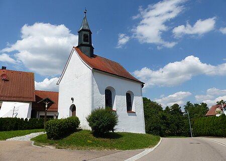 Kleine weiße Filialkirche mit rotem Dach und kleinem Turm oben drauf an einer Straße bei blauem Himmel mit Wolken. Umgeben von grüner Wiese und Hecken, daneben ein Jesuskreuz aus Holz. Im Hintergrund weiteres weißes Haus zu sehen.