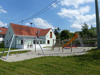 Spielplatz mit einer Schaukel, Klettergerüst, einem Sandkasten mit roter Umrandung und oranger Rutsche, vor einem türkisfarbenen Haus mit rotem Dach, bei blauem Himmel. Umgeben von einer grünen Wiese und Bäumen mit Kieselsteine an den Spielgeräten.