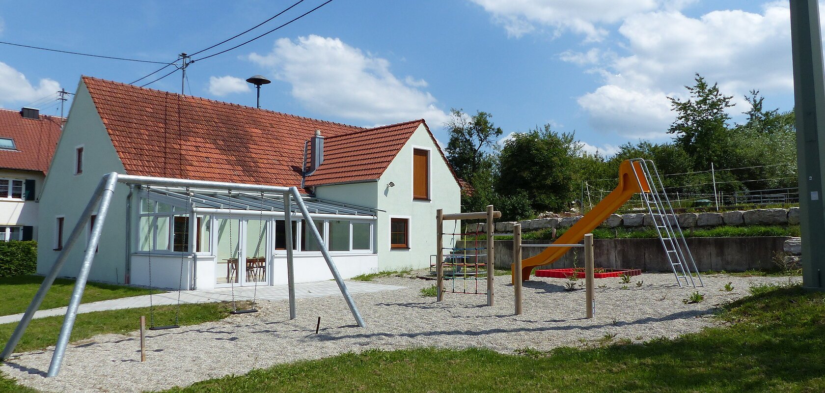 Spielplatz mit einer Schaukel, Klettergerüst, einem Sandkasten mit roter Umrandung und oranger Rutsche, vor einem türkisfarbenen Haus mit rotem Dach, bei blauem Himmel. Umgeben von einer grünen Wiese und Bäumen mit Kieselsteine an den Spielgeräten.